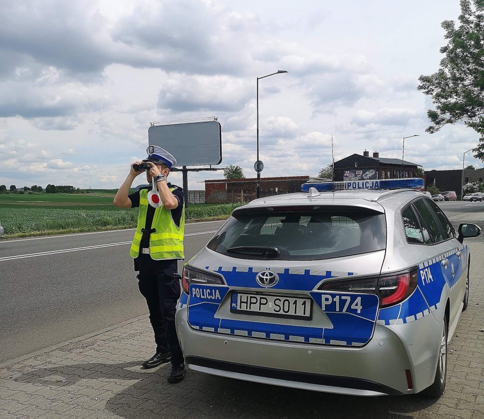 policjant na drodze przy radiowozie patrzy przez lornetkę