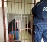 podejrzany siedzi w pomieszczeniu okratowanym, obok stoi policjant