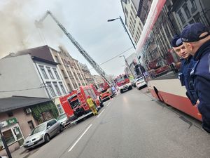 policjanci stoją na ulicy, w tle pojazdy straży pożarnej, zwyżka i płonący dach budynku