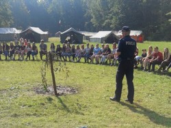 zdjęcie kolorowe policjant w czasie prelekcji na obozie harcerskim dzieci i młodzież siedzą na ławkach