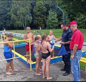 na zdjęciu policjant i ratownik wodny rozdaje dzieciom na basenie gadżety profilaktyczne