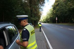 na zdjeciu policjant wykonuje kontrolę pojazdu, za nim stoi policjant