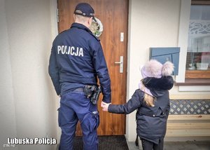 na zdjęciu policjant trzyma dziecko za rękę wchodzi do mieszkania