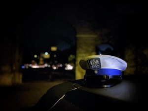 Zdjęcie przedstawia policyjną czapkę, która położona jest na dachu radiowozu. W tle widoczny jest cmentarz.