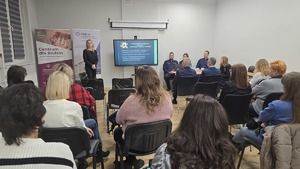 Zdjęcie przedstawia siedzących uczestników debaty oraz w drugim tle mówcę - psycholog ośrodka interwencji kryzysowej