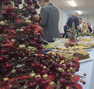 Zdjęcie przedstawia choinkę dekoracyjną oraz stół zastawiony potrawami. W ich tle widnieją policjanci i osoby cywilne.