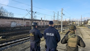 Zdjęcie przedstawia trzech funkcjonariuszy - Policji, Straży Ochrony Kolei oraz żołnierza Wojska Polskiego na tle torowiska kolejowego