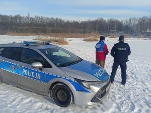 Zdjęcie przedstawia radiowóz Policji, za którym stoi ratownik wraz z policjantem. W tle znajduje się zamarznięty akwen wodny