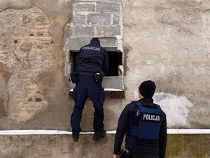 Zdjęcie przedstawia dwóch policjantów, którzy kontrolują opuszczony budynek przez dziurę w ścianie.