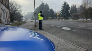 Zdjęcie przedstawia policyjny radiowóz, a w jego tle umundurowanego funkcjonariusza Policji