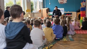 Zdjęcie przedstawia przedszkolaków, którzy słuchają policjanta w tle