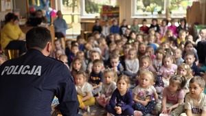 Zdjęcie przedstawia odwróconego policjanta, który skierowany jest w dzieci znajdujące się w tle