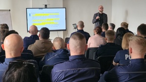 Na zdjęciu widnieją policjanci oraz pracownicy miejskiego ośrodka pomocy społecznej, a w tle widoczny jest jeden z prowadzących zajęcia, który omawia obowiązujące przepisy