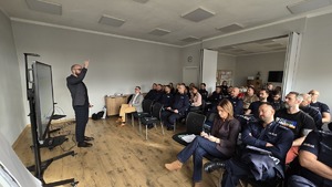 Zdjęcie przestawia siedzących pracowników miejskiego ośrodka pomocy społecznej wraz z policjantami, którzy słuchają prowadzącego spotkanie szkoleniowe.