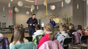 Na zdjęciu widnieją odwrócone plecami dzieci, które słuchają policjanta w tle