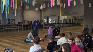 Zdjęcie ukazuje wpatrzone dzieci w ratownika Wodnego Ochotniczego Pogotowia Ratunkowego oraz policjanta, którzy pokazują jak prawidłowo udzielać pierwszej pomocy