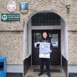 Na zdjęciu widnieje policjant trzymający plakat promujący bezpieczne spędzanie ferii zimowych przed wejściem do placówki oświatowej