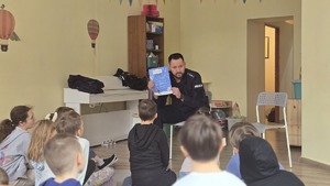 Na zdjęciu widnieją dzieci, które słuchają mundurowego omawiającego zasady policyjnego konkursu "Zimowy Dekalog"