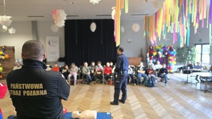 Na zdjęciu widnieje odwrócony strażak pożarny wraz z policjantem, na tle słuchających dzieci