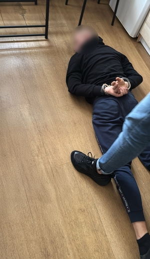 Zdjęcie przedstawia policjanta, który pilnuje zatrzymanego, leżącego na podłodze.