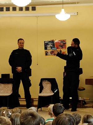 Zdjęcie przedstawia dzieci, które słuchają dzielnicowych w trakcie omawiania przez nich numerów alarmowych