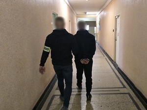 Na zdjęciu widoczny jest policjant, który prowadzi korytarzem osobę zatrzymaną