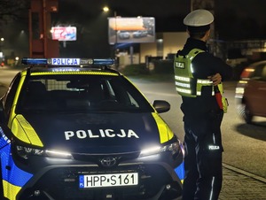 Na zdjęciu widoczny jest policjant stojący obok radiowozu. Mundurowy obserwuje ruch kołowy pojazdów