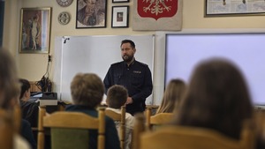 Na zdjęciu widać młodzież, która siedzi w ławkach, zwrócona w kierunku policjanta