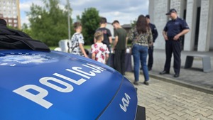 Zdjęcie przestawia zaparkowany, oznakowany radiowóz policji, a w tle widać stoisko profilaktyczne
