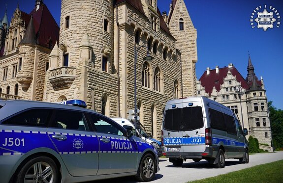 Na zdjęciu widoczne są oznakowane radiowozy policyjne, a w ich tle znajduje się Zamek Moszna