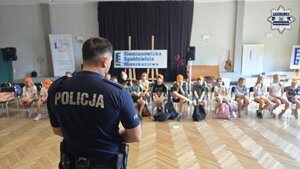 Zdjęcie przedstawia odwróconego policjanta w kierunku siedzących dzieci z półkolonii