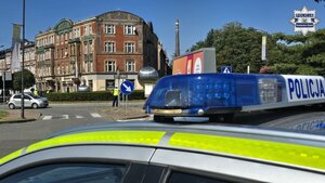 Zdjęcie przedstawia na pierwszym planie oznakowany radiowóz, a w jego tle policjantów drogówki