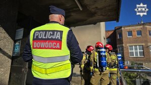 Zdjęcie przedstawia odwróconego plecami policjanta, a w jego tle widać strażaków pożarnych